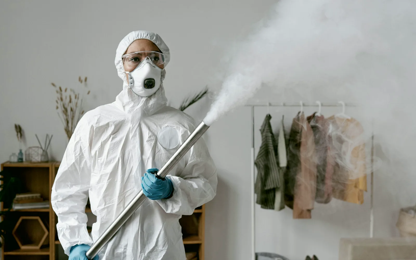 Técnico de dedetização com macacão branco, óculos e máscara segurando o aplicador de nebulização, com a névoa saindo em direção à arara de roupas
